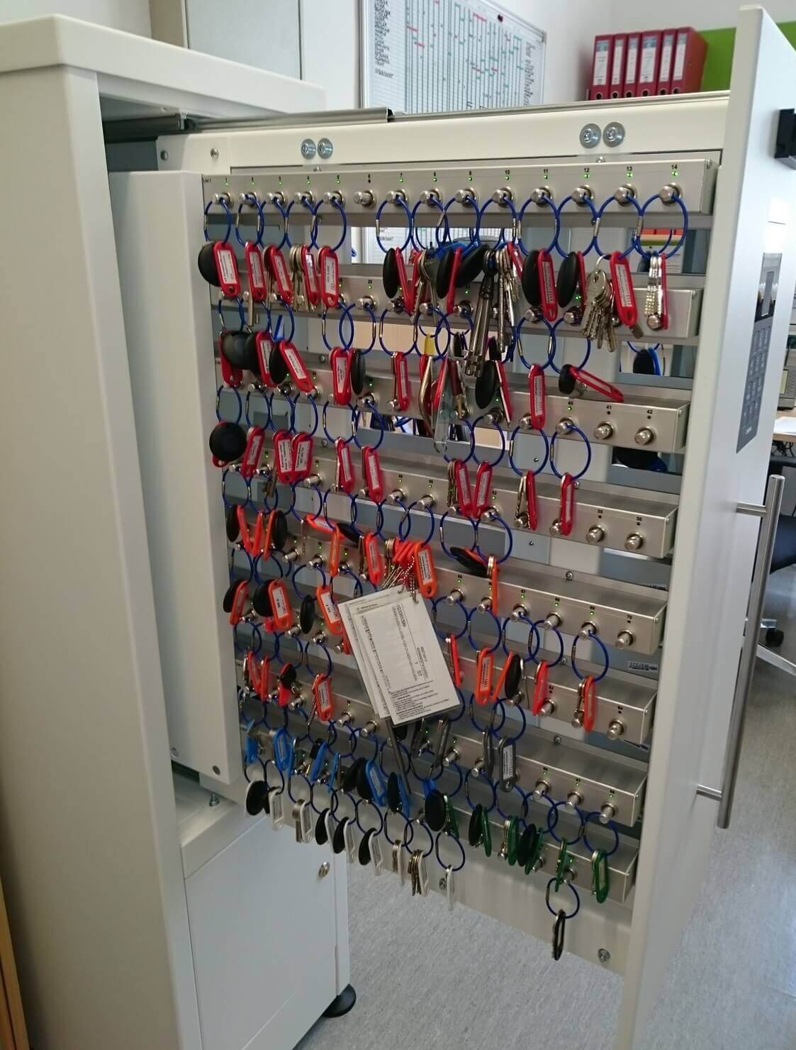 Offener Schlüsselschrank mit vielen Schlüsseln an farbigen Schlüsselanhängern und einer Inventarliste im Büro