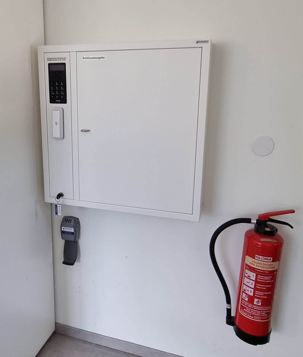 Weißer Schlüsselausgabe-Schrank mit elektronischem Codefeld und Griff, daneben ein roter Feuerlöscher an einer weißen Wand befestigt