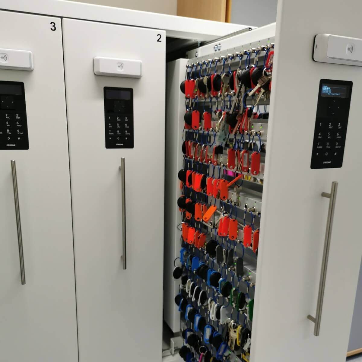 Geöffneter elektronischer Schlüsselschrank mit vielen sortierten Schlüsseln an farbigen Anhängern, daneben verschlossene Schränke mit Zahlenschlössern