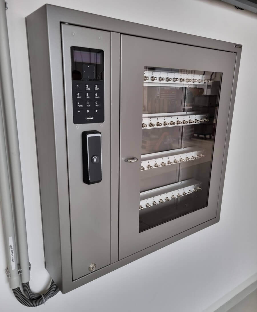 Metallischer Schlüsselschrank mit elektronischem Zugangssystem, Glastür und mehreren Haken zur Schlüsselaufbewahrung, an einer weißen Wand montiert