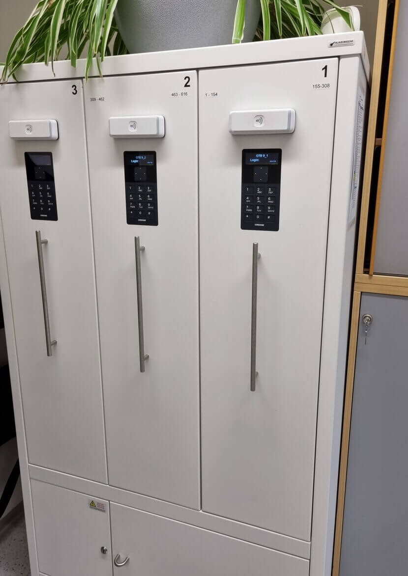 Drei weiße Schränke mit elektronischen Zahlenschlössern und langen Griffen, darauf steht ein grüner Blumentopf