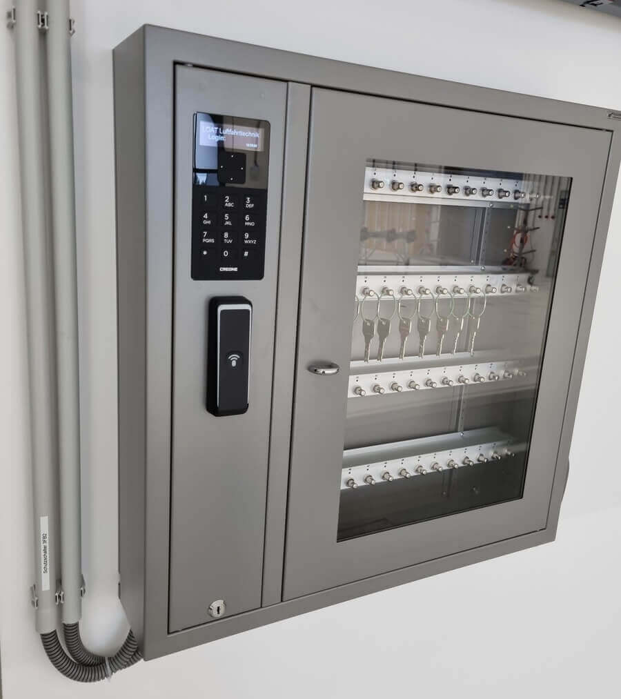 Grauer elektronischer Schlüsselkasten an der Wand mit Glastür, Zahlencode-Tastatur und RFID-Leser, darin mehrere Schlüssel sichtbar