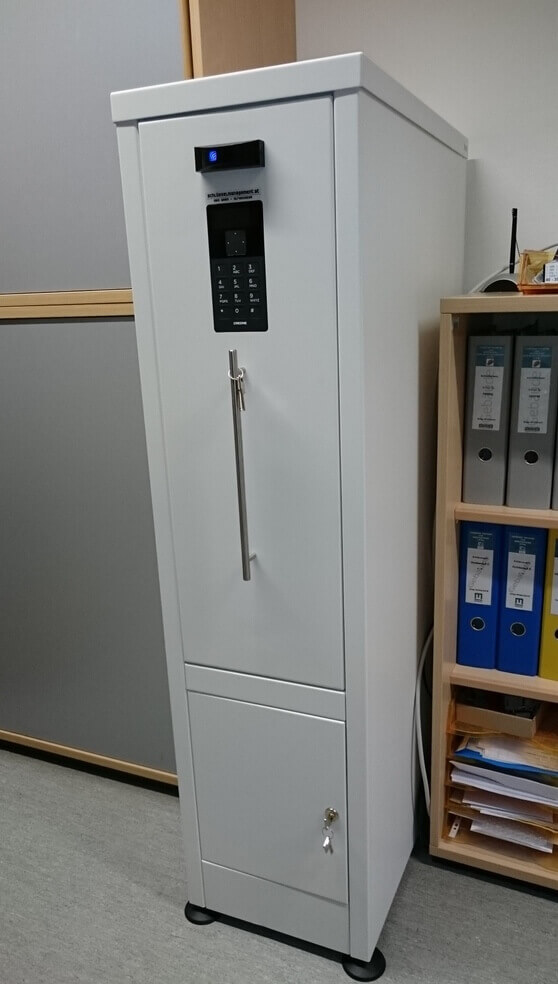 Weißer Waffenschrank mit elektronischem Zahlencode-Schloss und langer Griffstange, stehend in einem Büro neben einem Regal mit Aktenordnern