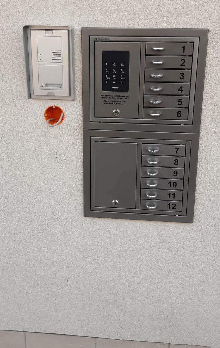 Briefkastenanlage aus Metall mit zwölf nummerierten Fächern, digitalem Zahlenschloss und Gegensprechanlage, montiert an einer weißen Wand