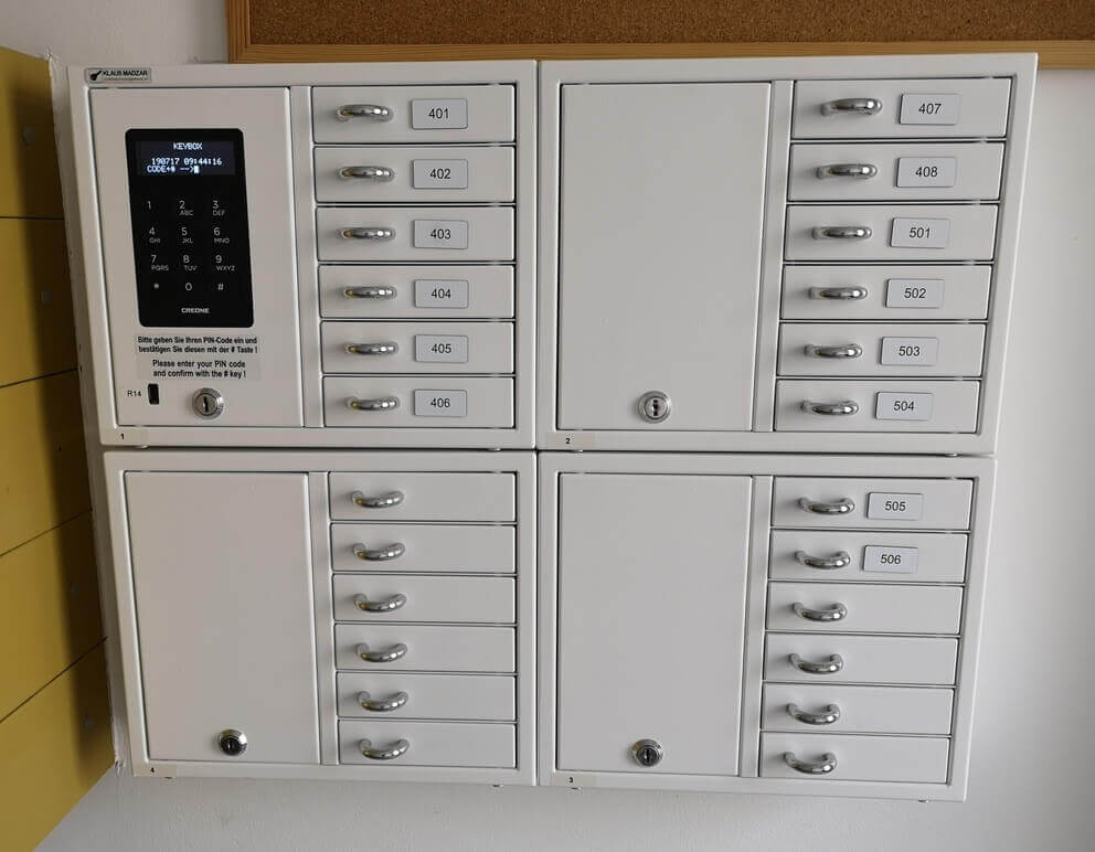 Weißer, wandmontierter Schlüsselkasten mit elektronischer PIN-Tastatur und insgesamt sechzehn nummerierten Fächern für die Schlüsselaufbewahrung.