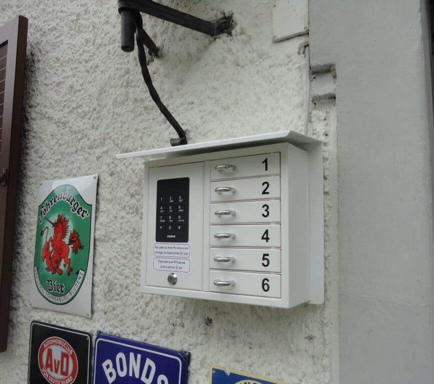Elektronischer Schlüsseltresor an einer Hauswand mit sechs nummerierten Fächern, daneben Vintage-Schilder
