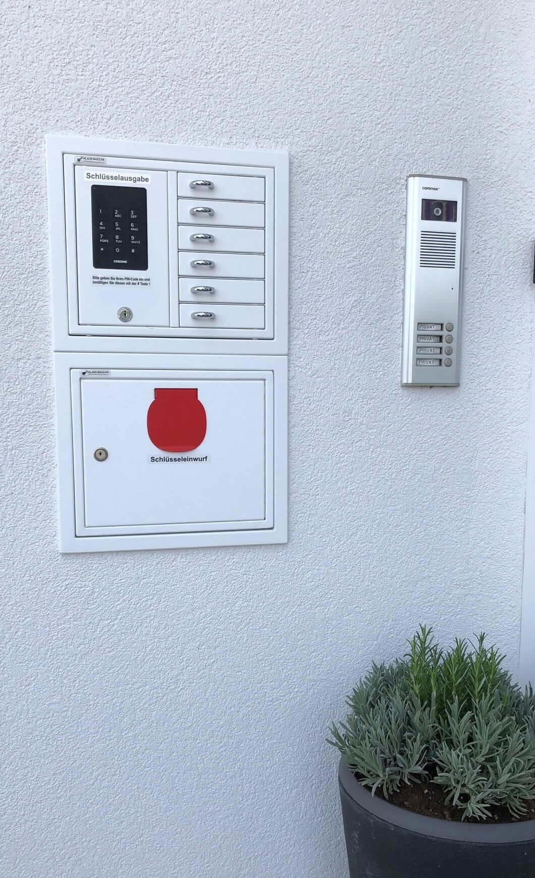 Elektronisches Schlüsselausgabesystem mit mehreren Fächern, Schlüsselrückgabefach sowie moderner Gegensprechanlage neben einem Pflanzkübel an einer weißen Wand