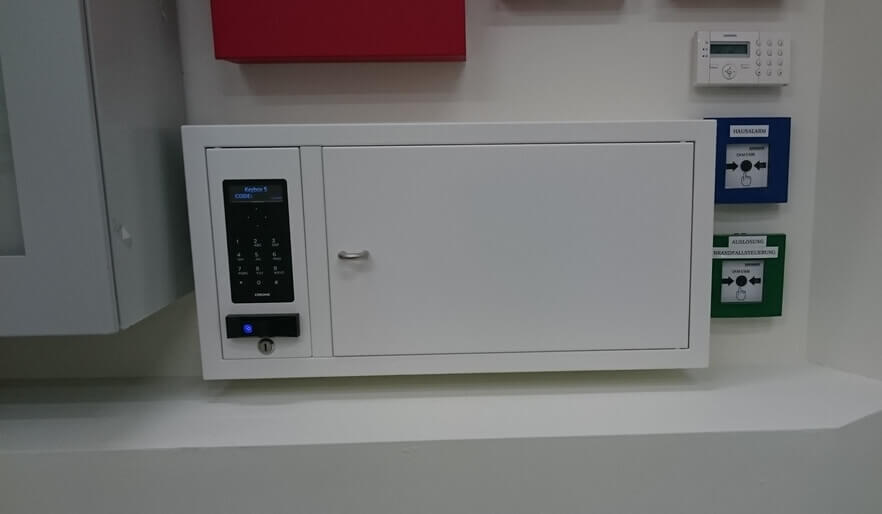 Weißer elektronischer Schlüsselschrank mit Tastatur zur Schlüsselverwaltung, montiert an einer Wand neben weiteren Sicherheitseinrichtungen